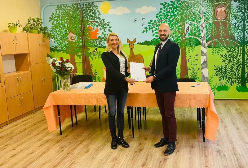 Zdjęcie w orientacji poziomej przedstawia dwie osoby stojące w sali przedszkolnej i trzymające wspólnie biały dokument. Kobieta po lewej stronie ma na sobie czarną marynarkę, dżinsy i czarne botki, natomiast mężczyzna po prawej jest ubrany w ciemną marynarkę, koszulę z krawatem oraz bordowe spodnie. Przed nimi znajduje się stół nakryty łososiowym obrusem, na którym stoi wazon z bukietem kwiatów. Tło stanowi kolorowy mural przedstawiający leśne zwierzęta, drzewa i ptaki na jasnym, błękitnym niebie, co sugeruje przyjazną przestrzeń edukacyjną. Po lewej stronie kadru widoczna jest jasna, drewniana zabudowa szafek, a całe wnętrze wykończone jest podłogą imitującą jasne drewno.