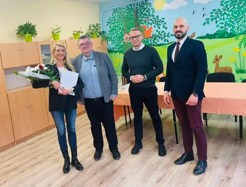 Zdjęcie wykonane w orientacji pionowej przedstawia grupę czterech osób stojących w jasnej sali przedszkolnej lub szkolnej. Po lewej stronie widoczna jest blondynka w czarnym żakiecie, trzymająca bukiet kwiatów oraz dokument, a obok niej stoi trzech mężczyzn w eleganckich strojach. Tło tworzy jasna ściana z kolorowym malowidłem ściennym przedstawiającym las, zwierzęta oraz drzewa, a także drewniane szafki i stół przykryty łososiowym obrusem. Kompozycja zdjęcia jest symetryczna, a postaci ustawione są w jednej linii frontem do obiektywu, co nadaje całości oficjalny charakter. Jasne oświetlenie i pastelowe barwy wnętrza sugerują przyjazne, edukacyjne otoczenie, idealne do publikacji relacji z wydarzeń społecznych lub uroczystości.