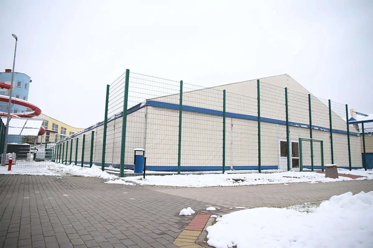 Zdjęcie w orientacji poziomej ukazuje nowoczesną halę sportową o konstrukcji namiotowej, otoczoną wysokim zielonym ogrodzeniem panelowym. Obiekt posiada jasne, kremowe poszycie z niebieskimi akcentami i znajduje się na terenie wyłożonym kostką brukową, częściowo pokrytą warstwą śniegu. W lewej części kadru widoczna jest czerwona rura zjeżdżalni wodnej należącej do sąsiedniego kompleksu oraz wysoka latarnia uliczna. Na pierwszym planie po lewej stronie ustawiony jest niebieski kosz na śmieci, a tło stanowi zachmurzone, jasne niebo. Całość kompozycji o proporcjach 3:2 przedstawia infrastrukturę sportową w otoczeniu zimowego krajobrazu.
