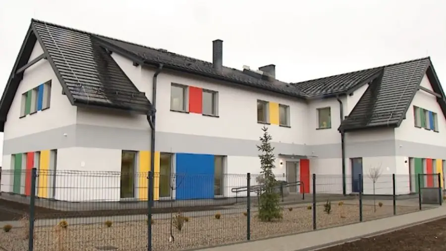 Zdjęcie przedstawia nowoczesny budynek użyteczności publicznej lub przedszkole w układzie poziomym o proporcjach boków 16:9. Biała elewacja budynku została urozmaicona szarym pasem biegnącym wzdłuż ścian oraz wyrazistymi, wielokolorowymi akcentami w odcieniach czerwieni, niebieskiego, żółtego i zielonego wokół okien. Dwuspadowy dach pokryty jest ciemną dachówką, a całość posesji otoczona jest metalowym ogrodzeniem panelowym. Przed budynkiem znajduje się niewielki ogródek z młodymi nasadzeniami drzew iglastych oraz utwardzone chodniki. Architektura obiektu jest przejrzysta, a liczne przeszklenia w połączeniu z kolorowymi panelami nadają fasadzie przyjazny i estetyczny charakter.